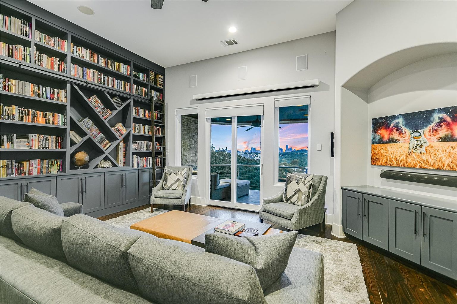 This is an interior shot of a living room featuring a large built-in bookcase filled with books, a gray sectional sofa, and two armchairs facing a sliding glass door that leads to a balcony with a city view. The room is decorated in neutral tones, with gray cabinetry and a modern aesthetic, creating a cozy and sophisticated atmosphere.