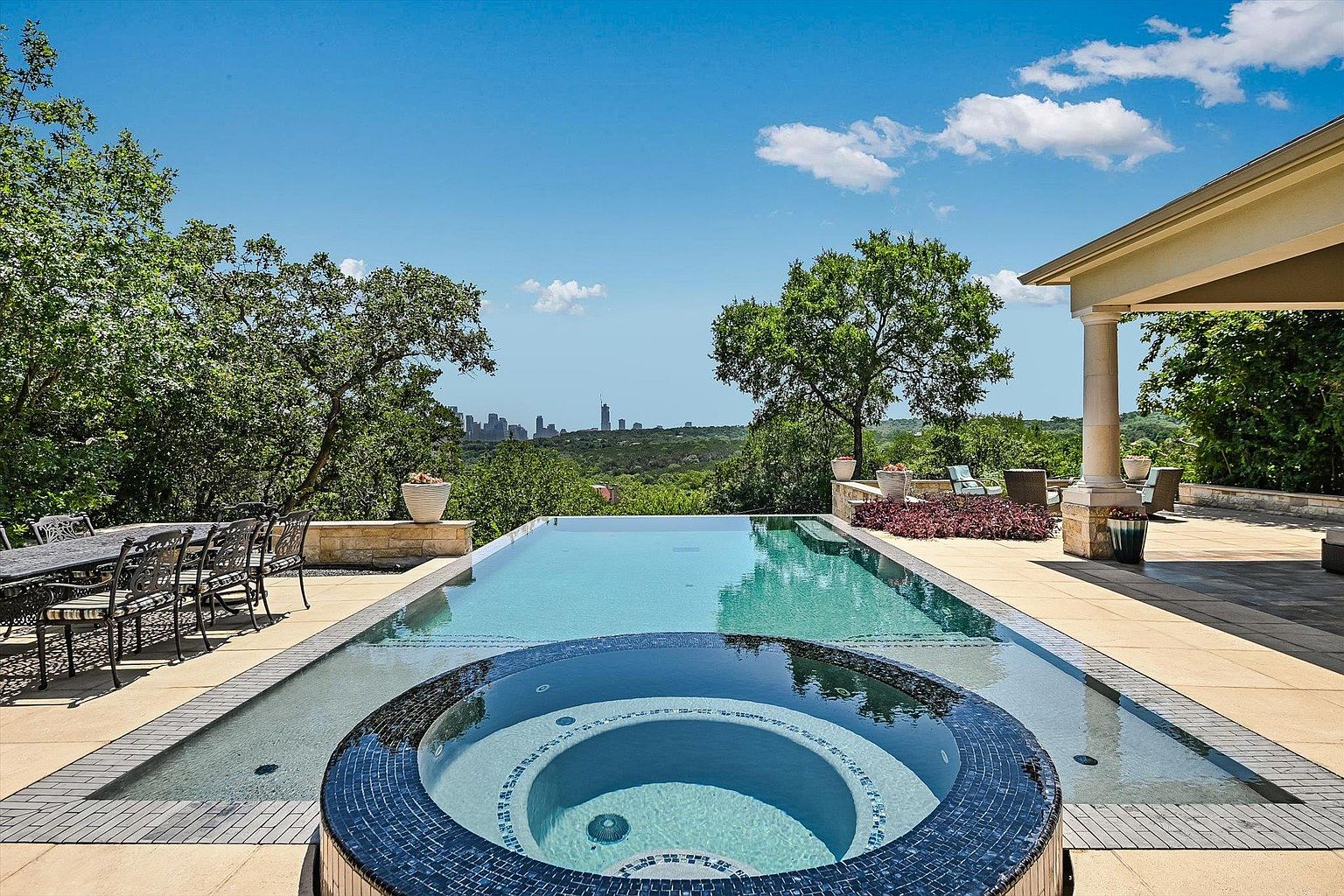 This image showcases a luxurious outdoor pool and spa area, featuring a modern infinity pool design with an integrated circular spa. The pool area is surrounded by a well-maintained patio, lush greenery, and mature trees, offering a serene and private setting. In the distance, a city skyline adds a touch of urban sophistication to the natural landscape.