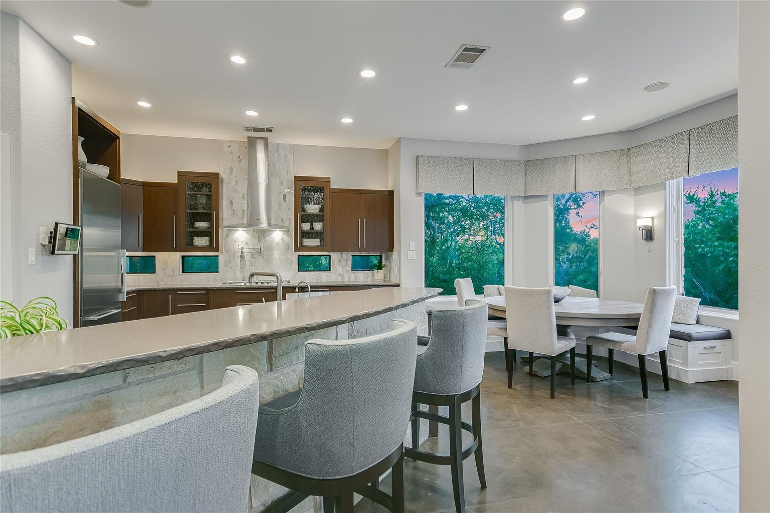 This is a well-lit, modern kitchen featuring dark wood cabinetry, stainless steel appliances, and a large island with a light-colored countertop. The kitchen seamlessly transitions into a dining area with a round table and comfortable seating, all bathed in natural light from the surrounding windows. The perspective is from the kitchen island, looking towards the dining area.