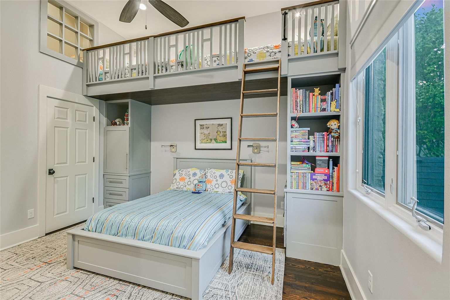 This is a charming bedroom featuring a unique loft bed design with built-in storage and a ladder for access. The room is decorated in a neutral color palette with pops of color in the bedding and accessories. A window provides natural light, and the overall impression is cozy and functional.
