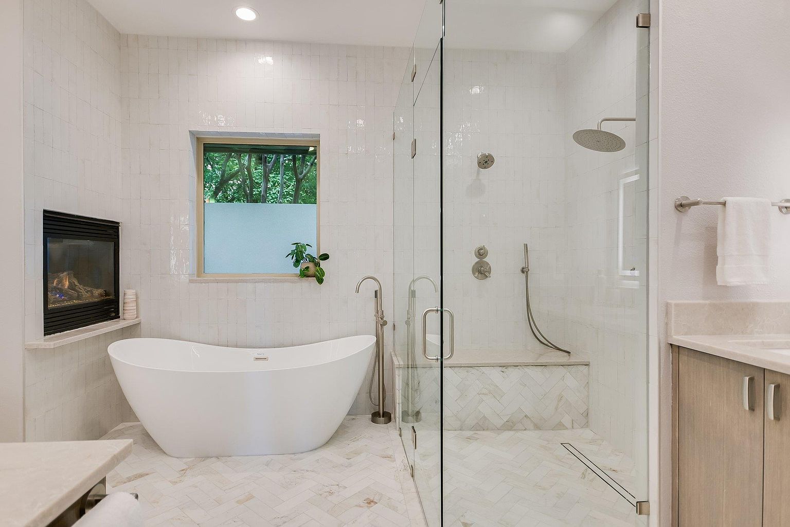 This is a luxurious primary bathroom featuring a freestanding white bathtub, a glass-enclosed shower with a built-in bench, and a modern fireplace. The walls and floors are adorned with light-colored tiles in a herringbone pattern, creating a bright and spacious feel. The room also includes a window with a frosted lower half for privacy and a vanity with light wood cabinetry.