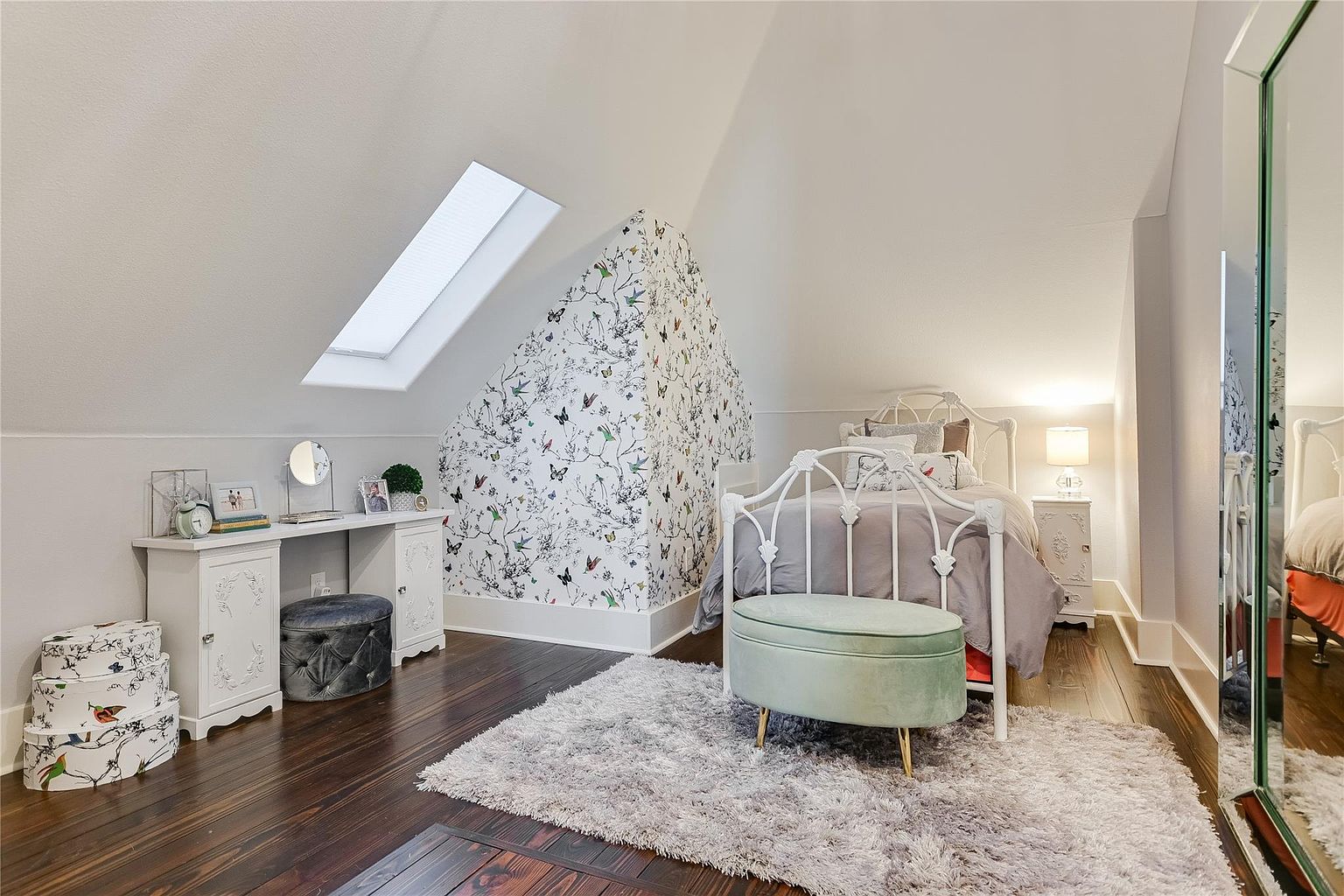 This is a charming bedroom featuring a white iron bed frame, a plush gray rug, and a unique wallpaper accent wall with butterfly and floral patterns. The room includes a white vanity with decorative details and a skylight that brightens the space. The overall impression is cozy and stylish, perfect for a guest room.