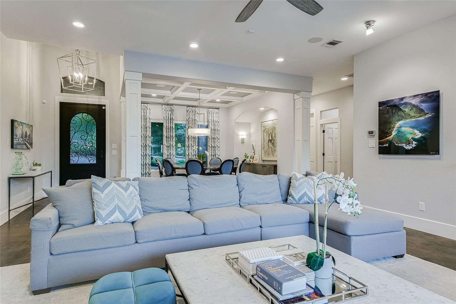 This interior shot showcases a spacious living room with a large, comfortable sectional sofa in a light gray hue. The room is well-lit with recessed lighting and features a decorative ceiling design that visually connects it to the adjacent dining area. A marble-topped coffee table and stylish decor add to the room's inviting and sophisticated ambiance.