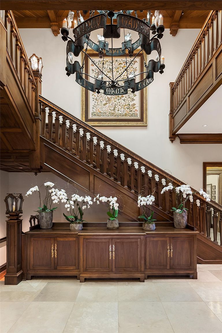 This grand entryway features a sweeping wooden staircase with intricate balusters, serving as the focal point of the space. A large, ornate metal chandelier hangs from the ceiling above a console table adorned with five potted white orchids. The neutral stone flooring and warm wood tones create an elegant, traditional atmosphere, while a framed piece of art adds a decorative touch to the wall.