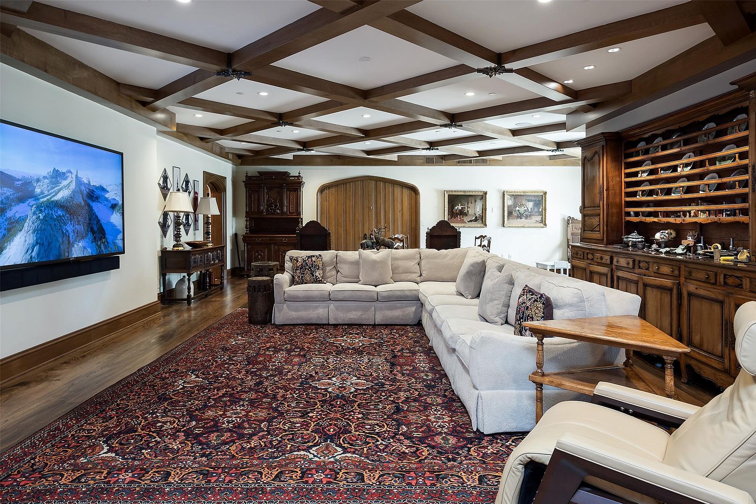 This spacious living room features a coffered wood-beam ceiling and a large, plush sectional sofa arranged on a traditional patterned area rug. The room is anchored by a large wall-mounted television on one side and an extensive, built-in wooden hutch with open shelving on the other. The space exudes a classic, refined atmosphere with warm wood tones and traditional decor elements.