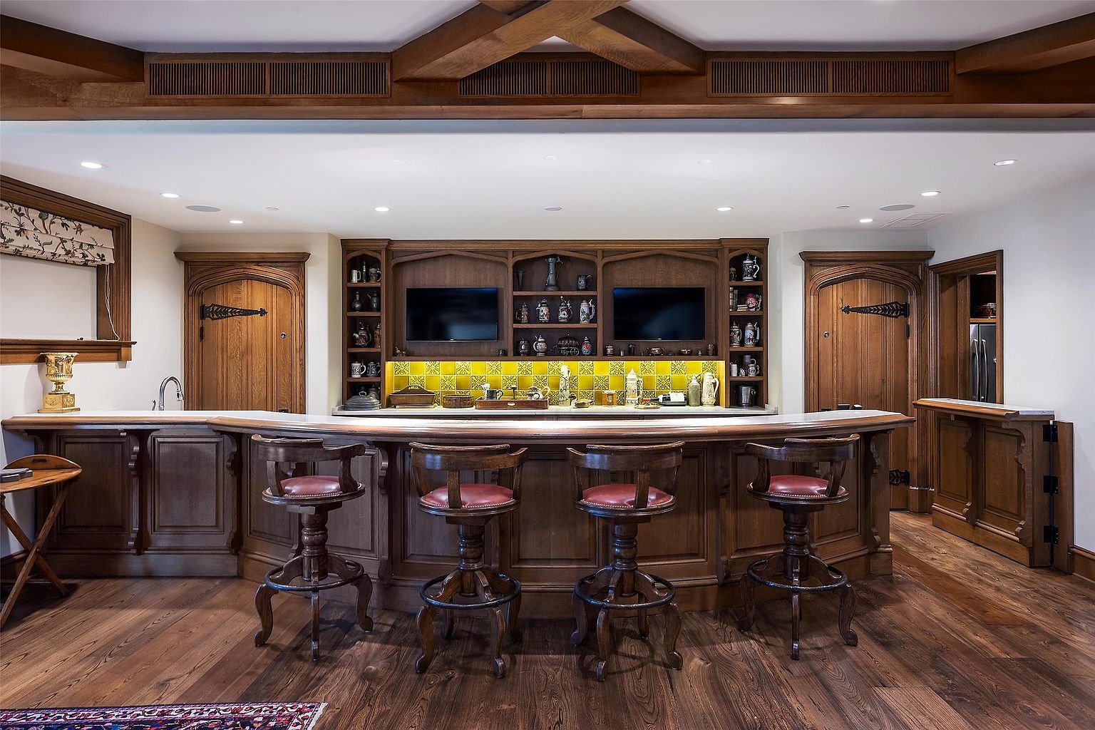 This sophisticated home bar area features rich, dark wood cabinetry and a curved bar counter with four matching stools topped with red leather seats. The back bar is accented by a vibrant yellow tiled backsplash and integrated shelving displaying a collection of steins and glassware, flanked by two televisions. The space is framed by arched wooden doorways and exposed ceiling beams, creating a warm, pub-like atmosphere perfect for entertaining.
