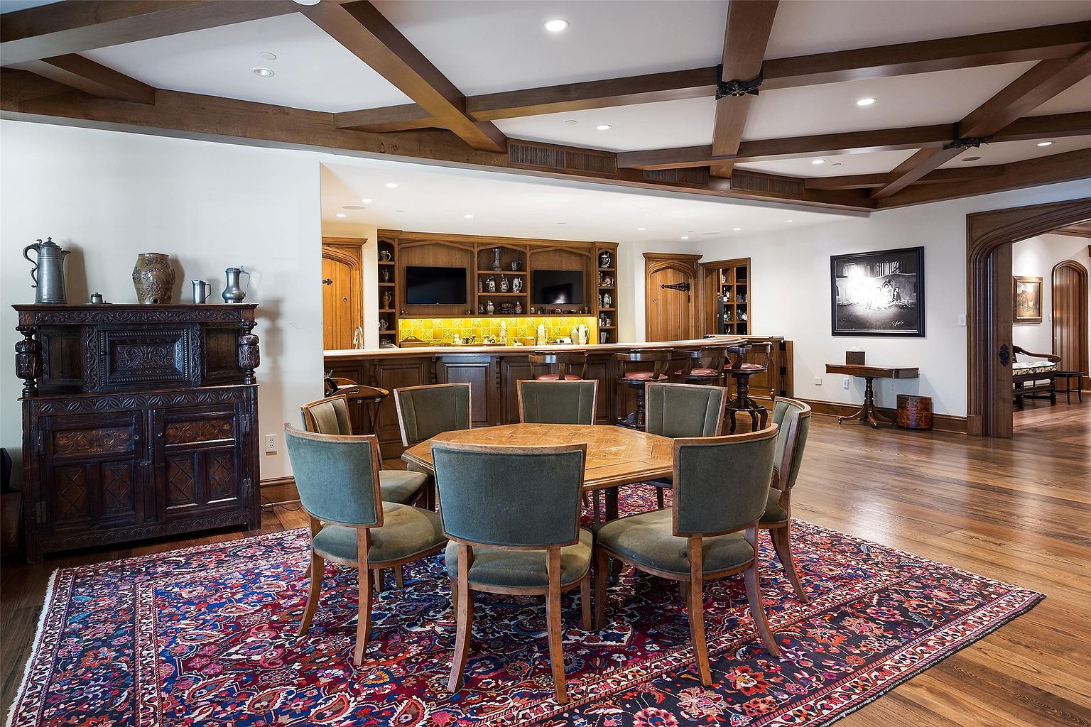 This spacious lower-level entertainment area features a large, custom-built wooden bar with integrated shelving and a yellow-tiled backsplash. In the foreground, a hexagonal wooden game table is surrounded by six upholstered chairs, all set atop a large, intricately patterned area rug. The room is defined by dark wood ceiling beams and warm, recessed lighting, creating a sophisticated and inviting atmosphere for hosting.