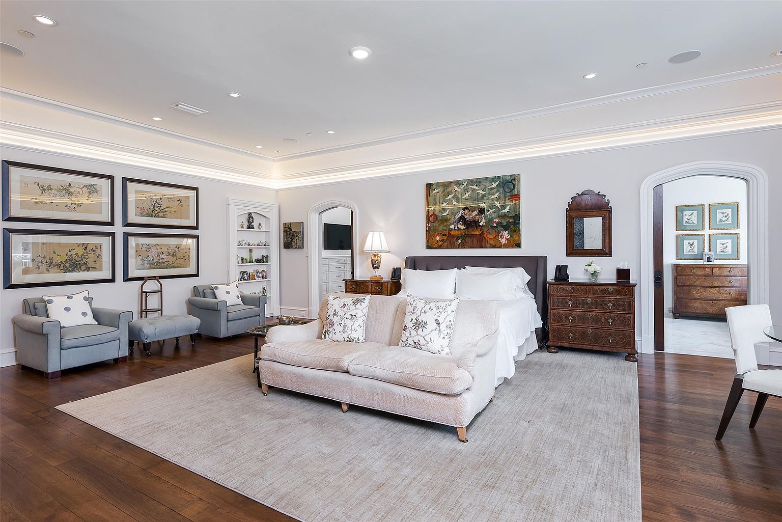 This spacious primary bedroom features a sophisticated design with a neutral color palette, dark wood flooring, and a large area rug anchoring the seating area. The room is furnished with a plush sofa at the foot of the bed, two armchairs, and an antique-style chest of drawers, all complemented by elegant wall art and recessed lighting. The perspective is wide and centered, showcasing the room's generous proportions and refined, traditional aesthetic.