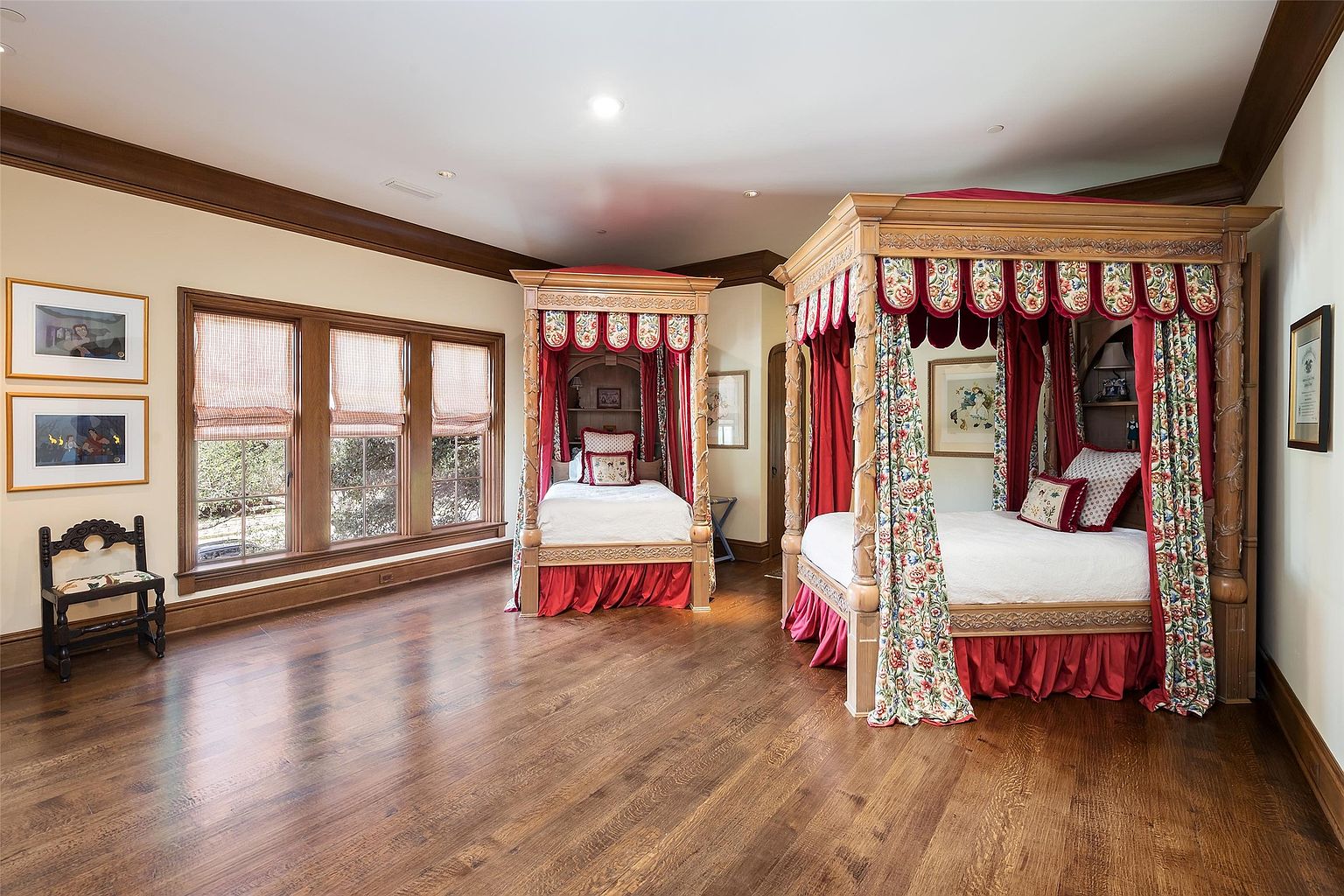 This spacious bedroom features two ornate, four-poster canopy beds with elaborate red and floral fabric drapery. The room is anchored by rich, dark wood flooring and crown molding, complemented by a large bank of windows with matching patterned shades. The traditional, opulent aesthetic is further enhanced by framed artwork on the walls and a small wooden chair, creating a formal and classic atmosphere.
