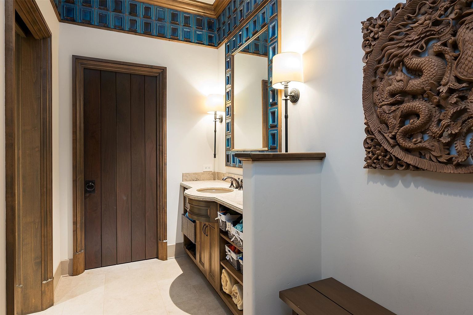 This bathroom features a unique vanity area with a stone countertop and open shelving for storage. The space is accented by a decorative blue tile border near the ceiling and a large, intricate wood carving mounted on the wall. A wooden bench sits in the foreground, and the room is illuminated by two wall-mounted sconces flanking a mirror.