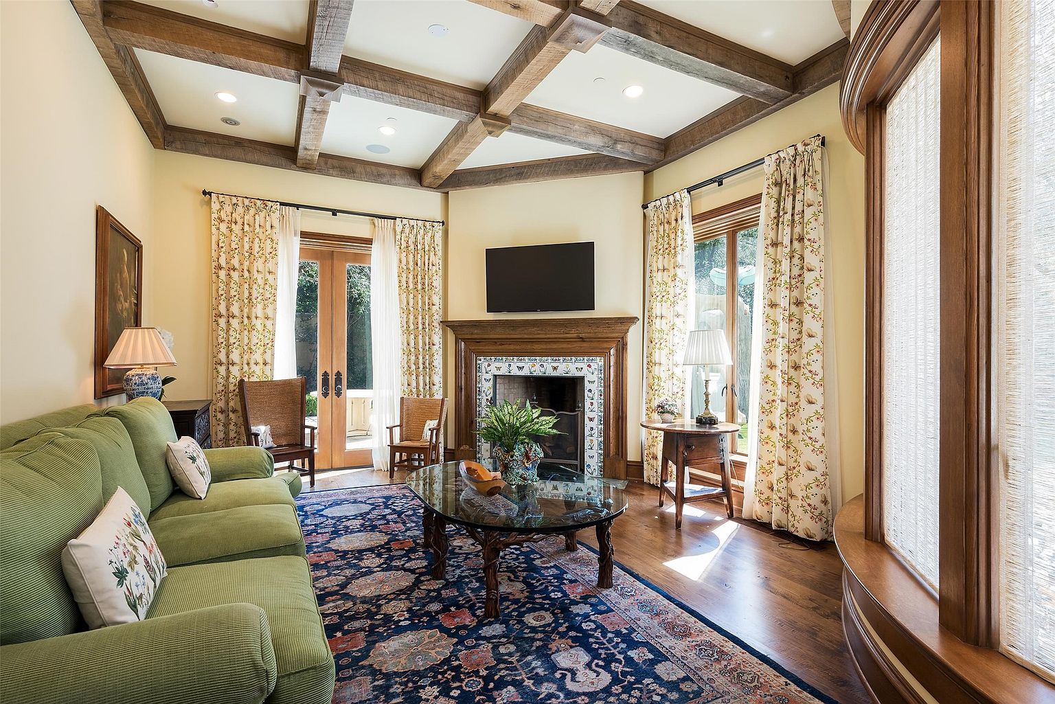 This inviting living room features a coffered ceiling with exposed wood beams and a central fireplace with a decorative tile surround. A green sofa sits atop a large, ornate area rug, while a glass-topped coffee table and two wooden chairs provide comfortable seating. The room is bathed in natural light from large windows and glass doors draped in floral-patterned curtains, creating a warm and traditional atmosphere.