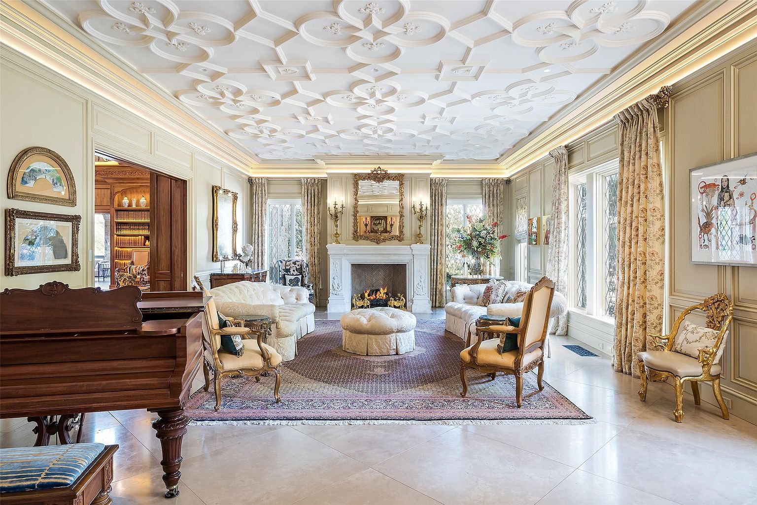 This elegant formal living room features a highly detailed, ornate coffered ceiling and a central fireplace with a decorative mantel and mirror. The space is furnished with plush, light-colored sofas, a central ottoman, and classic gold-accented armchairs arranged on a large, patterned area rug. Large windows draped in floral curtains allow natural light to fill the room, which also includes a grand piano in the foreground and a glimpse into an adjacent wood-paneled library.