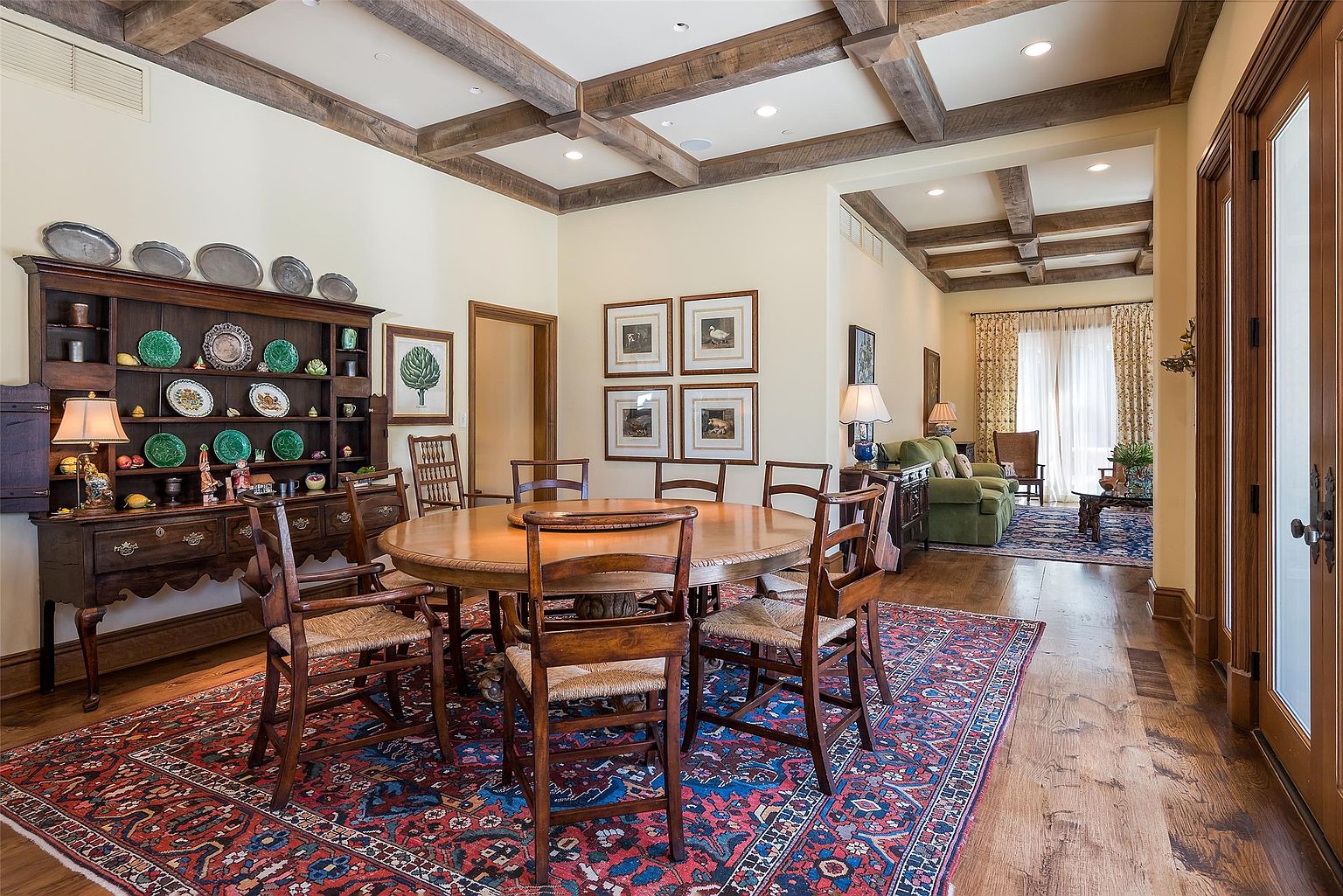 This elegant dining room features a large round wooden table surrounded by six chairs with woven seats, set atop a vibrant, intricately patterned area rug. A substantial dark wood hutch displays a collection of decorative plates and dishware, while the room is framed by a coffered ceiling with exposed wood beams. The perspective looks through the dining area into an adjacent living space, creating an open and inviting flow between the rooms.