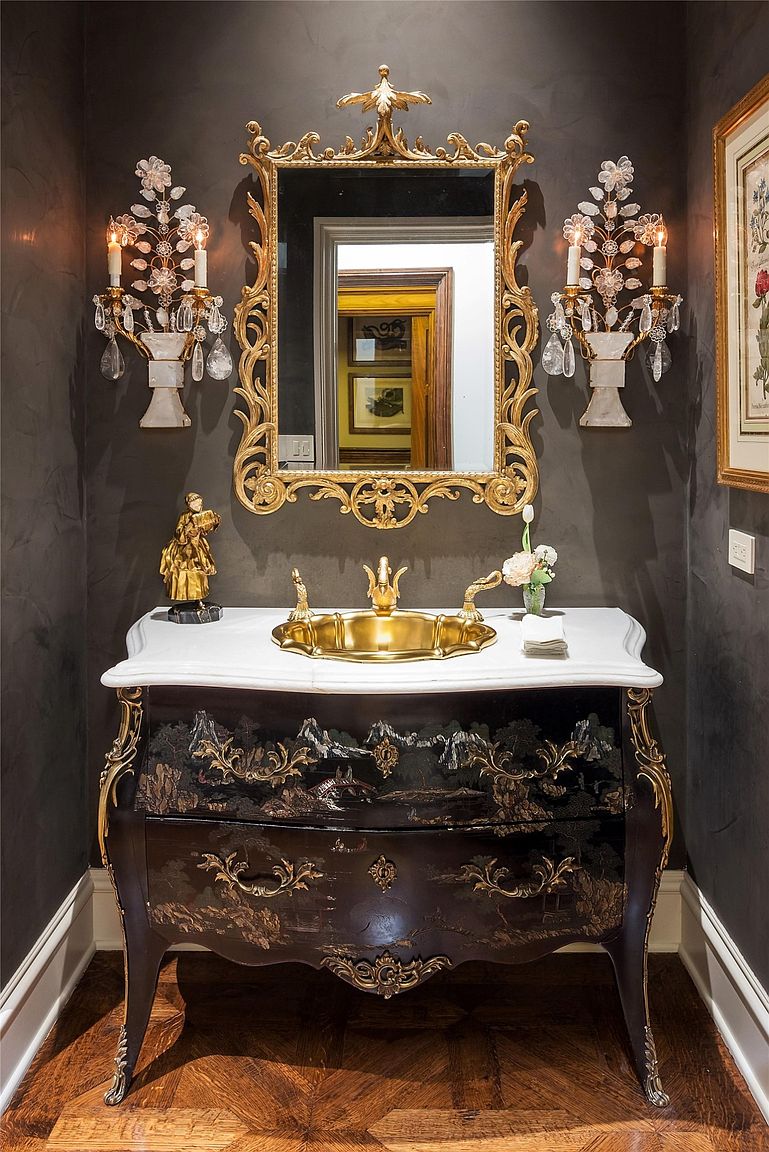 This elegant powder room features a sophisticated vanity with a dark, ornate cabinet base adorned with intricate landscape detailing and gold accents. A polished gold sink is set into a white stone countertop, complemented by a matching ornate gold-framed mirror and crystal-adorned wall sconces. The dark, textured walls create a dramatic backdrop, enhancing the luxurious and classical aesthetic of the space.