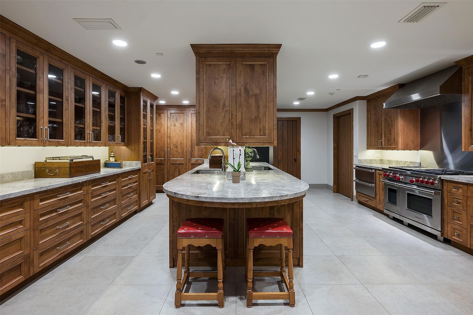 This spacious, traditional-style kitchen features extensive warm-toned wood cabinetry and a large central island topped with a light-colored stone countertop. The island includes a built-in sink and seating for two, while the perimeter is equipped with professional-grade stainless steel appliances, including a large range with a matching hood. The room is illuminated by recessed ceiling lights, creating a bright and functional workspace with a symmetrical layout.