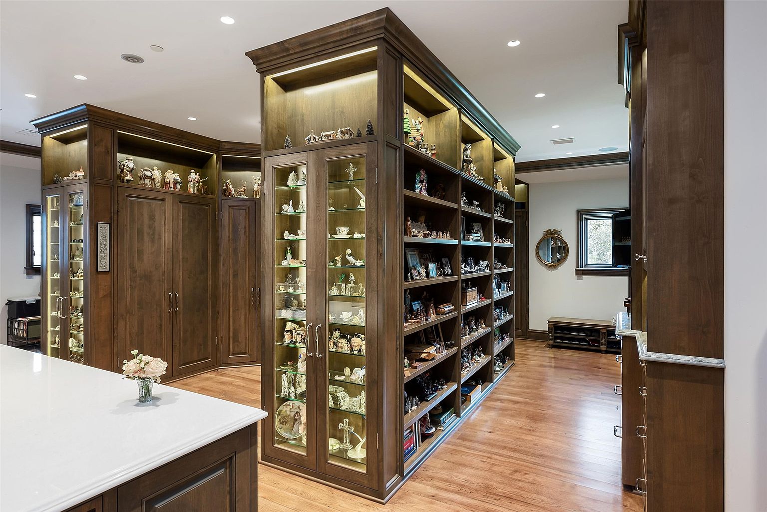 This interior space features a prominent, custom-built wooden display unit that serves as a central architectural divider, showcasing numerous collectibles on illuminated glass shelves. The room is finished with warm-toned wood flooring and recessed ceiling lighting, creating a sophisticated and gallery-like atmosphere. A white-topped island is visible in the foreground, while a small mirror and a bench are positioned near a window in the background, suggesting a transition area or a large, multi-purpose hallway.