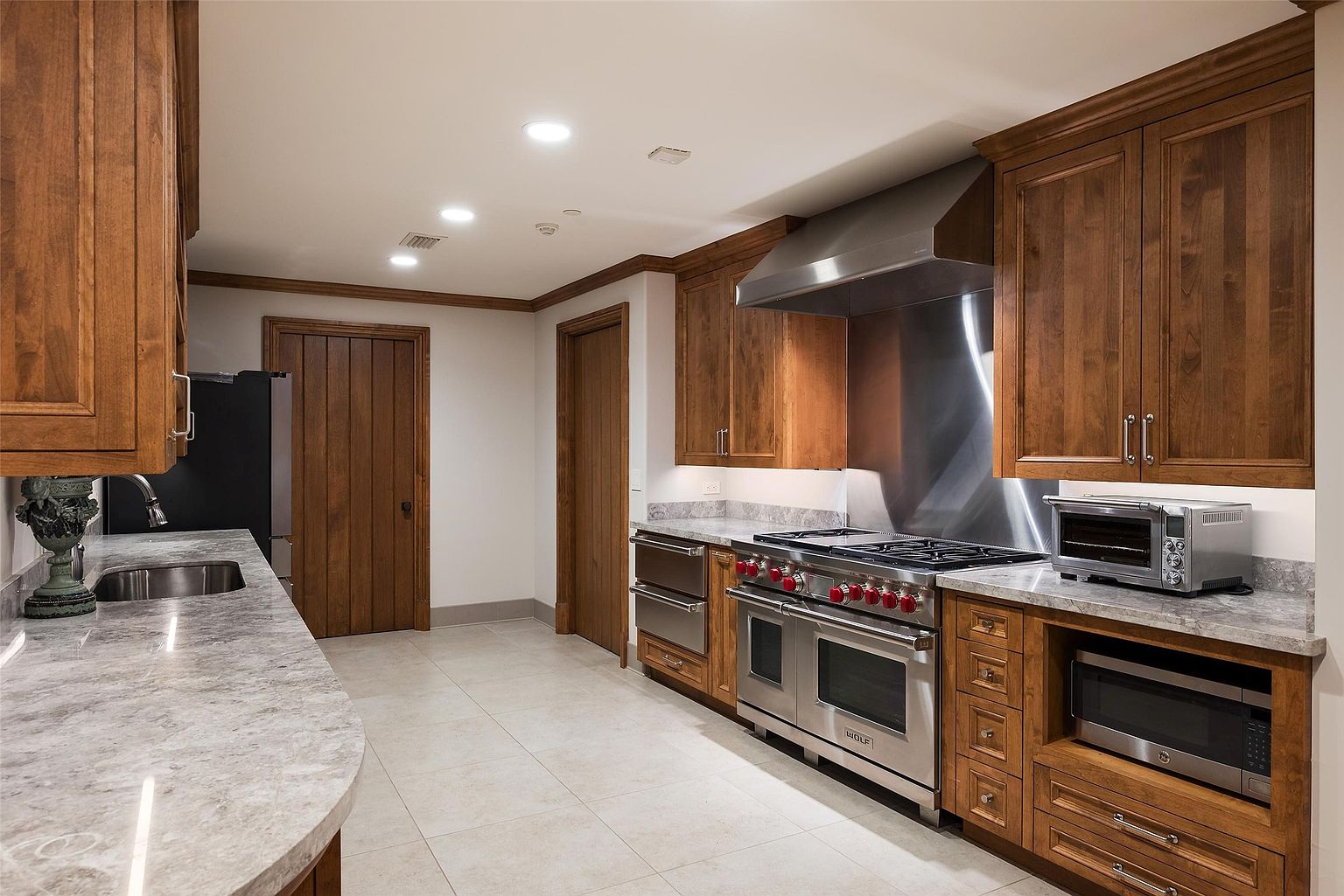 This kitchen features warm-toned wood cabinetry paired with light-colored stone countertops and a stainless steel backsplash behind a professional-grade range. The space is equipped with high-end appliances, including a large range with a matching vent hood, a built-in microwave, and a toaster oven. The perspective captures the length of the counter space and the transition toward the adjacent doorways, highlighting a clean, functional, and well-lit culinary environment.