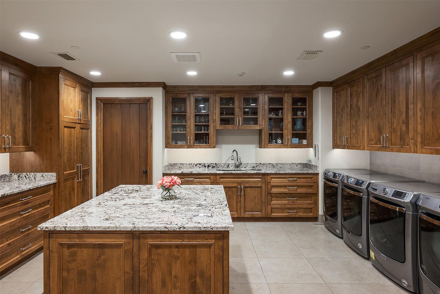 This spacious laundry room features warm-toned wood cabinetry, a large central island with a stone countertop, and a built-in sink area. The room is equipped with a side-by-side washer and dryer set, ample storage space, and recessed lighting throughout. The neutral tile flooring and clean lines create a functional and organized workspace.