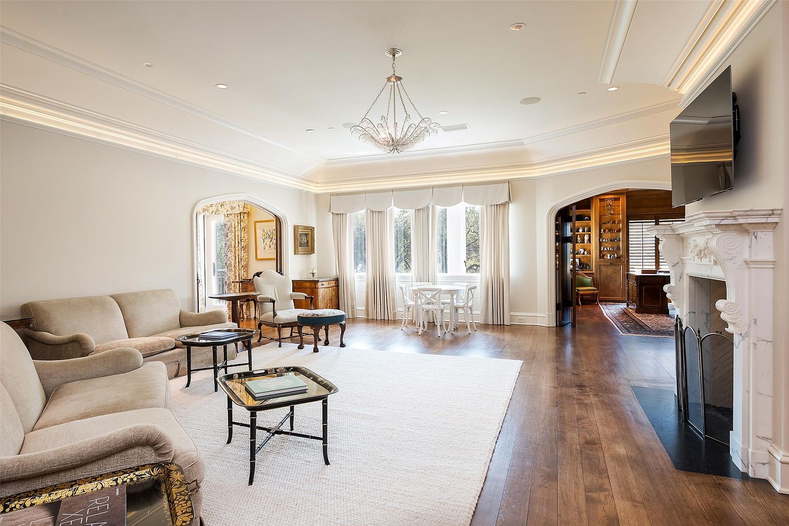 This elegant living room features a neutral color palette, hardwood flooring, and a large white area rug that anchors the seating area. A prominent fireplace with a decorative mantel serves as a focal point, while a crystal chandelier hangs from the tray ceiling, adding a touch of sophistication. The space is bright and airy, with large windows dressed in soft drapery and arched doorways leading to adjacent rooms, including a study visible in the background.