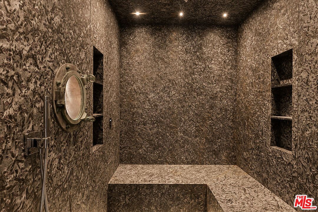 This unique shower space features walls and a built-in bench covered in a textured, dark, organic-patterned material. A circular, nautical-style porthole window is set into the left wall, accompanied by a modern handheld shower fixture. The space is illuminated by three recessed ceiling lights, creating a moody and immersive atmosphere.