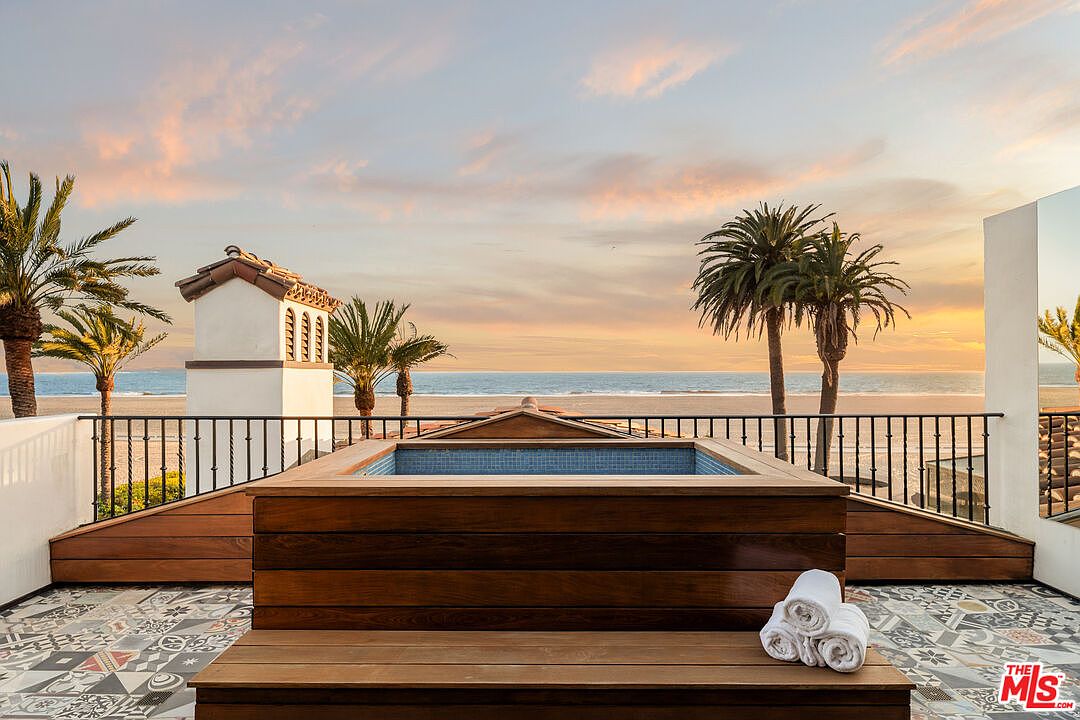This elevated deck features a prominent wood-encased spa overlooking a serene beach and ocean horizon at sunset. The space is finished with decorative patterned tile flooring and a black metal railing, creating a sophisticated outdoor retreat. The composition captures a wide-angle perspective that emphasizes the seamless transition between the private terrace and the expansive coastal view.