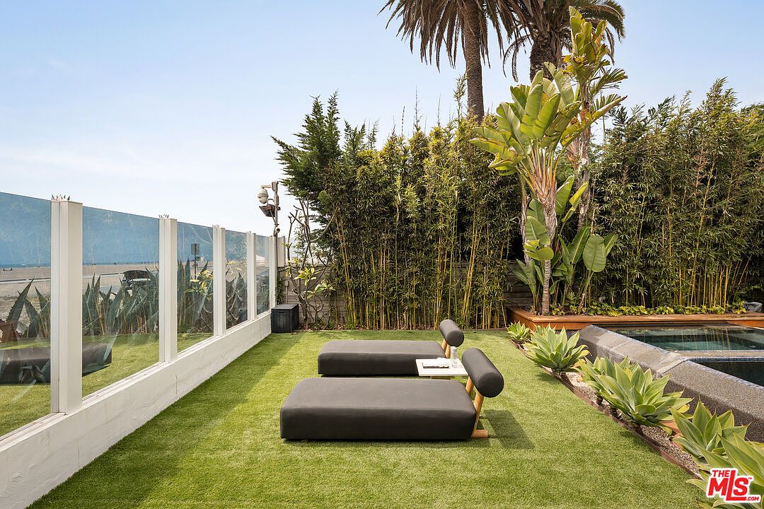 This serene outdoor space features a manicured artificial lawn furnished with two modern, low-profile lounge chairs. To the right, a small pool is bordered by lush agave plants and a tall bamboo privacy screen, while a glass railing on the left provides an unobstructed view of the beach beyond. The perspective is a wide, eye-level shot that captures the seamless blend of private luxury and coastal scenery.