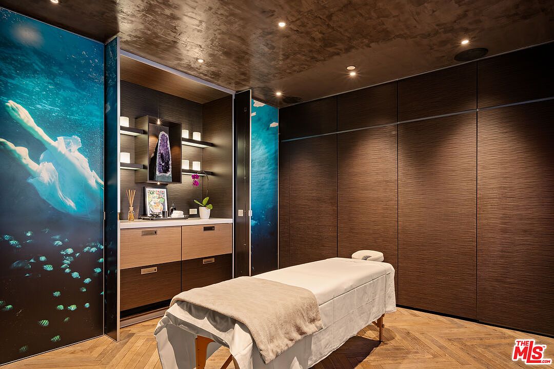 This image depicts a luxurious, spa-inspired massage room featuring a professional treatment table centered in the space. The room is accented by striking, illuminated wall panels depicting an underwater scene with a swimmer, contrasting with dark, textured wood-paneled walls and a herringbone-patterned wood floor. A built-in vanity area with open shelving and a decorative crystal display adds a sophisticated, tranquil touch to the professional wellness environment.