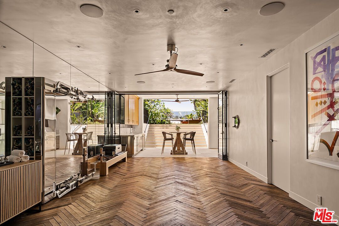 This interior space functions as a home gym, featuring a prominent Pilates reformer machine and a mirrored wall that enhances the sense of depth. The room is finished with rich, dark wood flooring laid in a classic herringbone pattern and opens directly to an outdoor patio through wide, folding glass doors. The perspective captures a seamless transition between the indoor workout area and the sunlit exterior dining space, creating an airy and sophisticated atmosphere.