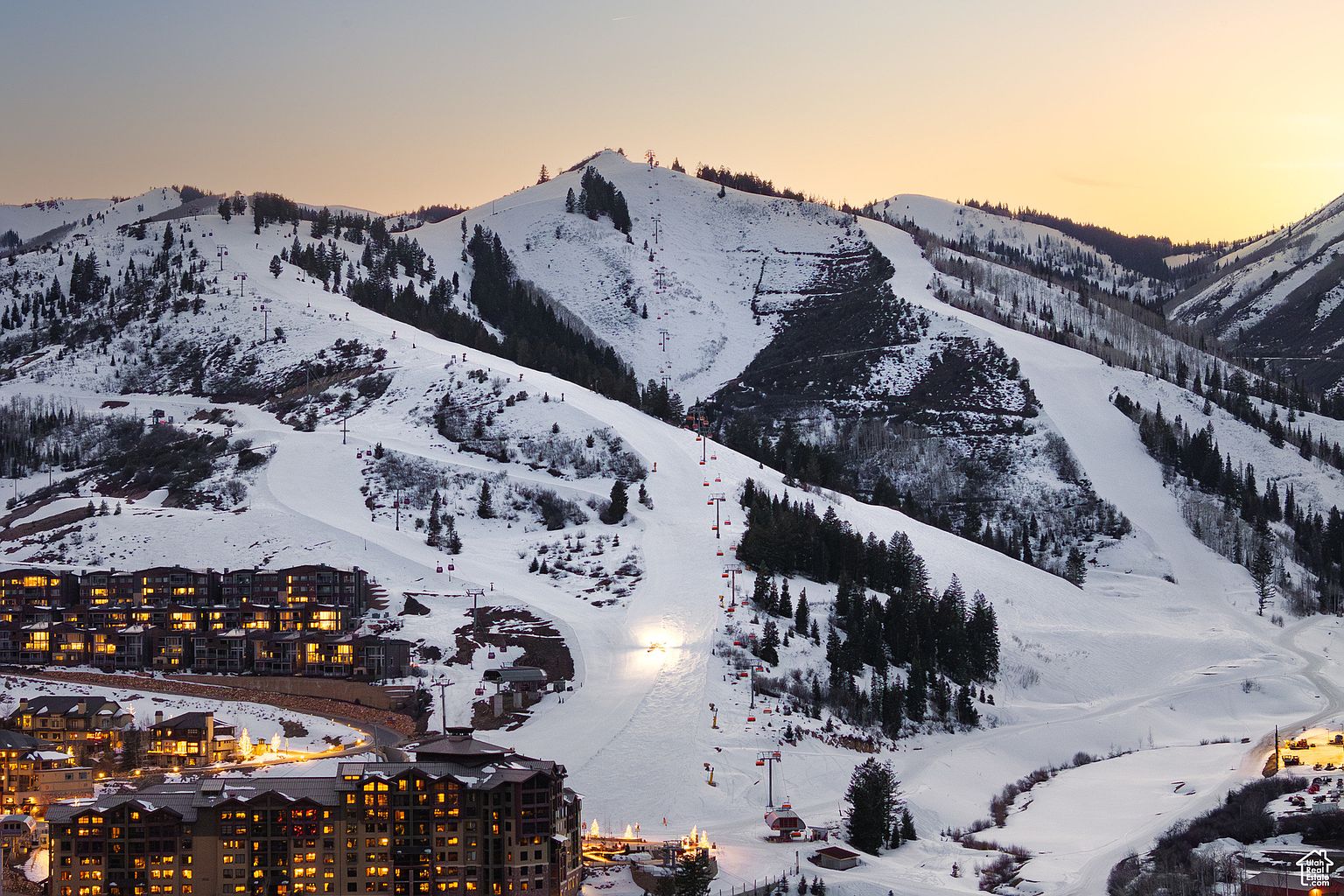 This aerial view showcases a ski resort at dusk, with snow-covered slopes and chairlifts ascending the mountain. Luxurious residential buildings are nestled at the base, their windows glowing warmly against the cool tones of the snow. The scene evokes a sense of winter luxury and recreational opportunity.