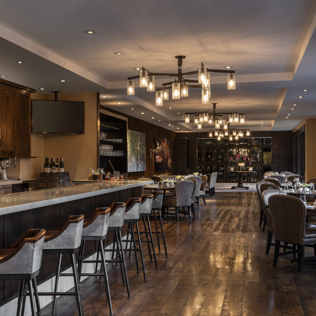 This image showcases a well-appointed dining room, potentially within a restaurant or high-end residential property. The space features dark wood flooring, elegant seating at multiple tables, and a bar area. Contemporary chandeliers add a modern touch, creating an inviting and sophisticated atmosphere.