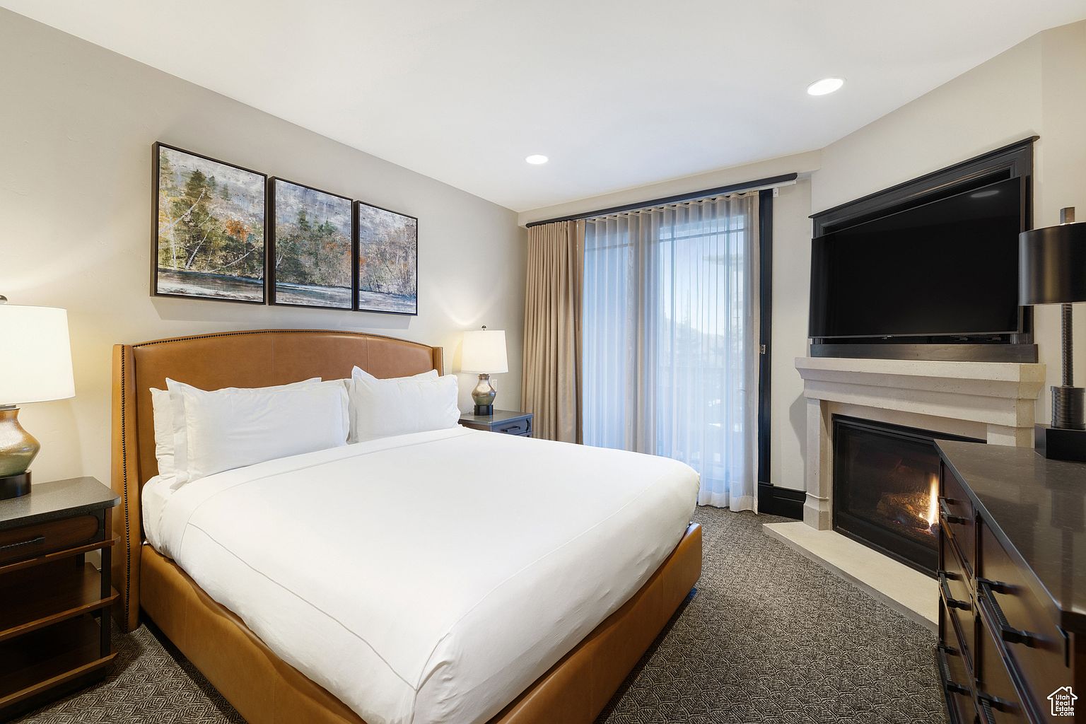 This is a well-appointed primary bedroom featuring a king-size bed with a leather headboard and crisp white linens. The room includes a fireplace with a mounted television above, creating a cozy and luxurious atmosphere. Artwork adorns the walls, and the room is illuminated by bedside lamps, enhancing the inviting ambiance.