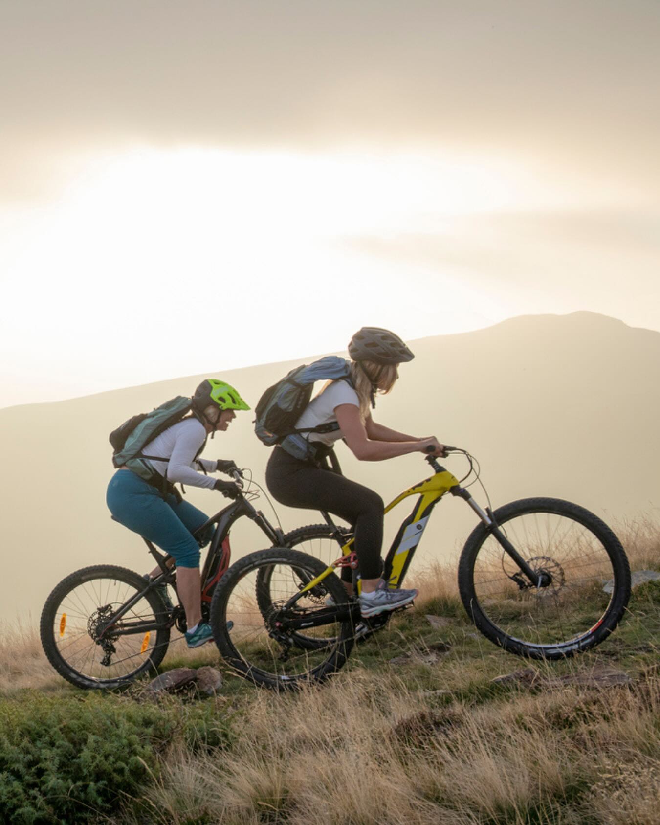 Two cyclists wearing helmets are riding their mountain bikes on a grassy ridge. The backdrop features mountains partially veiled by mist or fog illuminated by sunlight, which creates a sense of vastness. The scene suggests an active lifestyle and appreciation for nature.