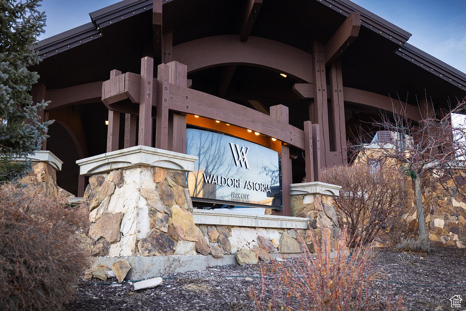 The image showcases the entryway of the Waldorf Astoria Park City, featuring a stone facade and a prominent sign. The architectural design includes dark wood beams and a well-lit sign displaying the hotel's name. The surrounding landscaping adds a touch of natural beauty to the entrance.