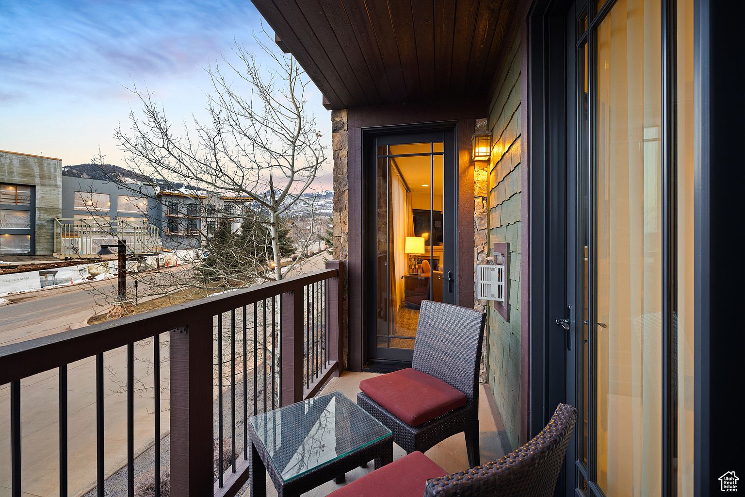 This image showcases a charming balcony area, furnished with a glass-topped table and two wicker chairs with red cushions. The balcony overlooks a street with modern buildings and bare trees, offering a pleasant outdoor space. The balcony features a dark wood railing and a door leading into the interior, suggesting a cozy and inviting extension of the living space.