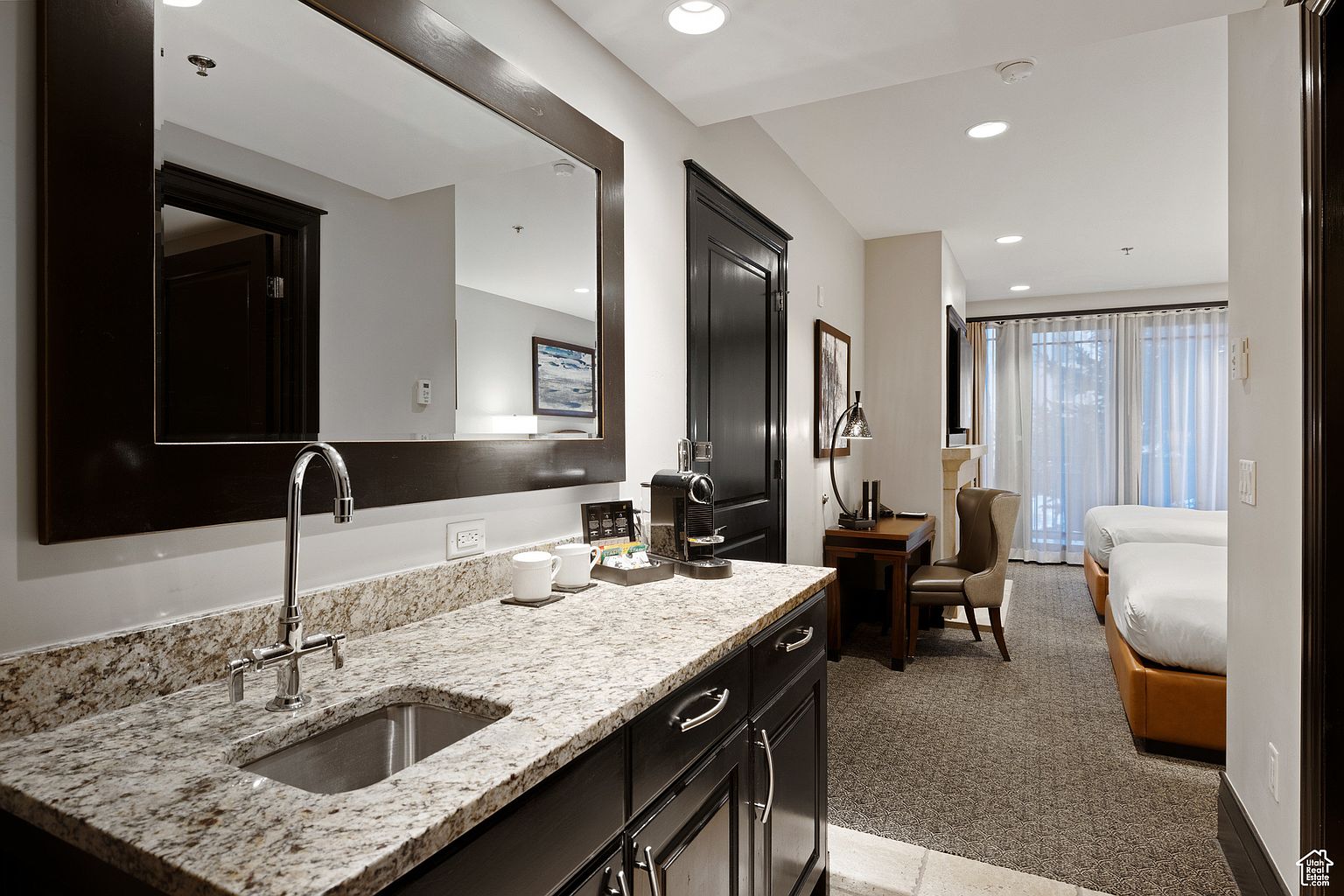 This interior shot showcases a hotel room or guest bedroom featuring a kitchenette area with a granite countertop, dark wood cabinetry, and a stainless steel sink. The room extends into a sleeping area with two beds, a desk, and a window with sheer curtains, creating a comfortable and inviting space. The overall impression is clean and well-maintained, suitable for a relaxing stay.