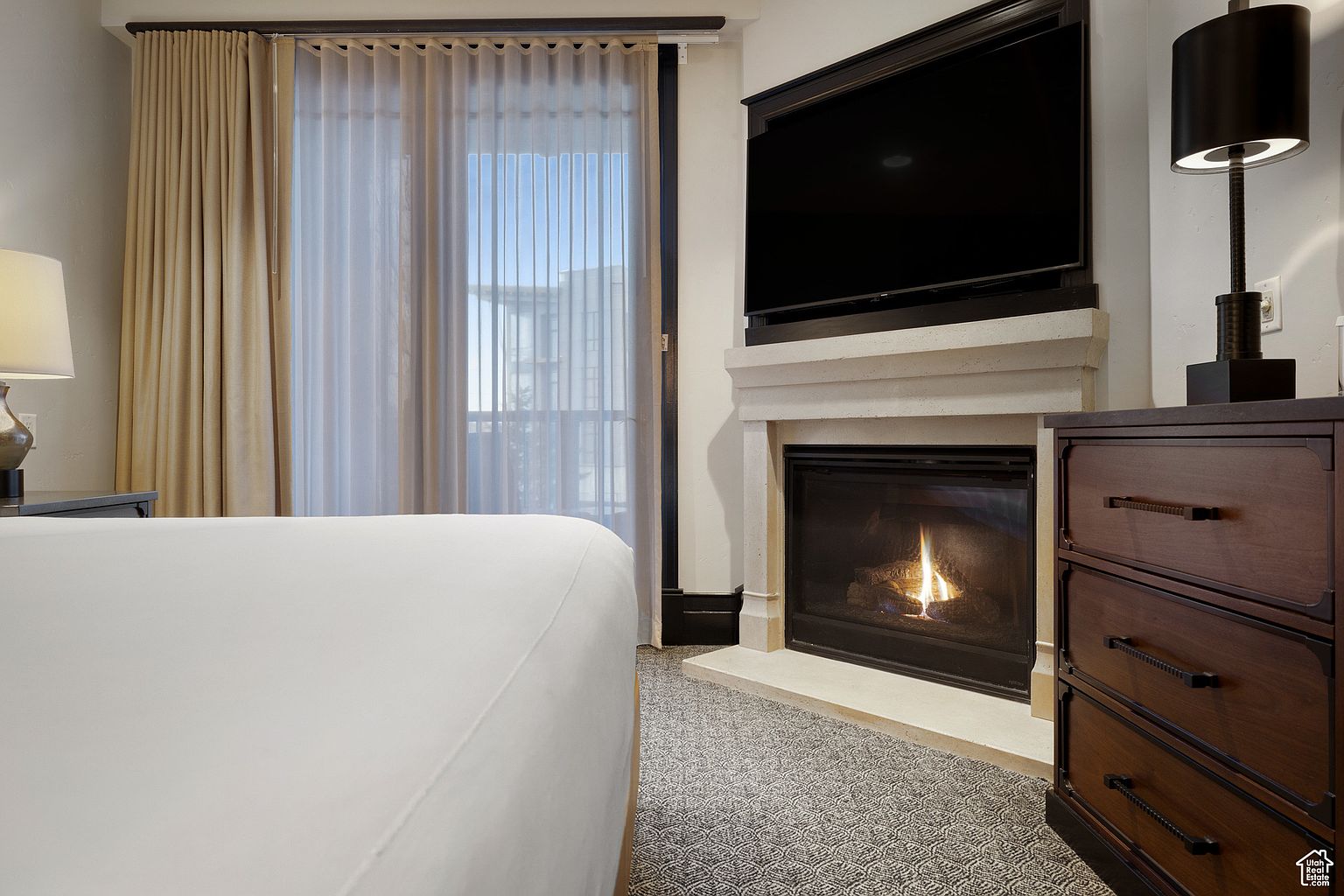 This is a cozy primary bedroom featuring a fireplace with a mounted television above it. A dark wood dresser sits to the right of the fireplace, topped with a modern lamp. The room has neutral-toned curtains and a patterned carpet, creating a warm and inviting atmosphere.