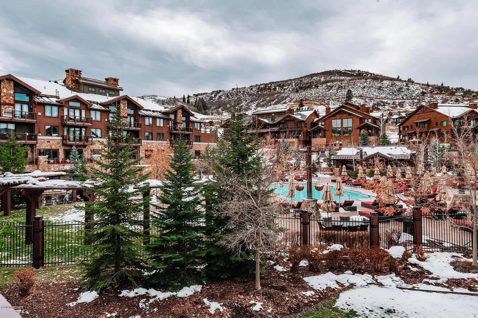 This image showcases a luxurious outdoor pool area at a mountain resort. Rustic wooden buildings with stone accents frame the scene, while snow-dusted landscaping and evergreen trees add a touch of winter charm. The pool is surrounded by lounge chairs and umbrellas, creating an inviting space for relaxation and recreation.