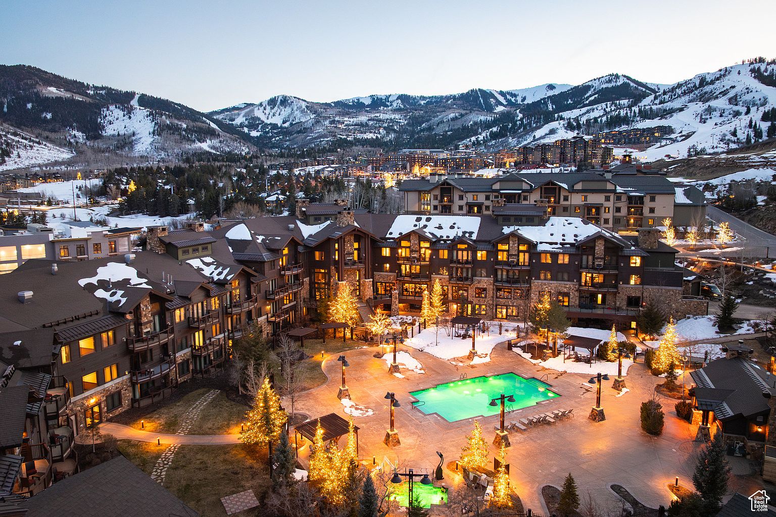 This aerial view showcases a luxurious resort nestled in a snowy mountain landscape. The property features multiple buildings with dark brown exteriors, illuminated windows, and snow-dusted roofs. A central courtyard includes a bright green swimming pool, decorated Christmas trees with lights, and inviting lounge areas, creating a warm and festive atmosphere.