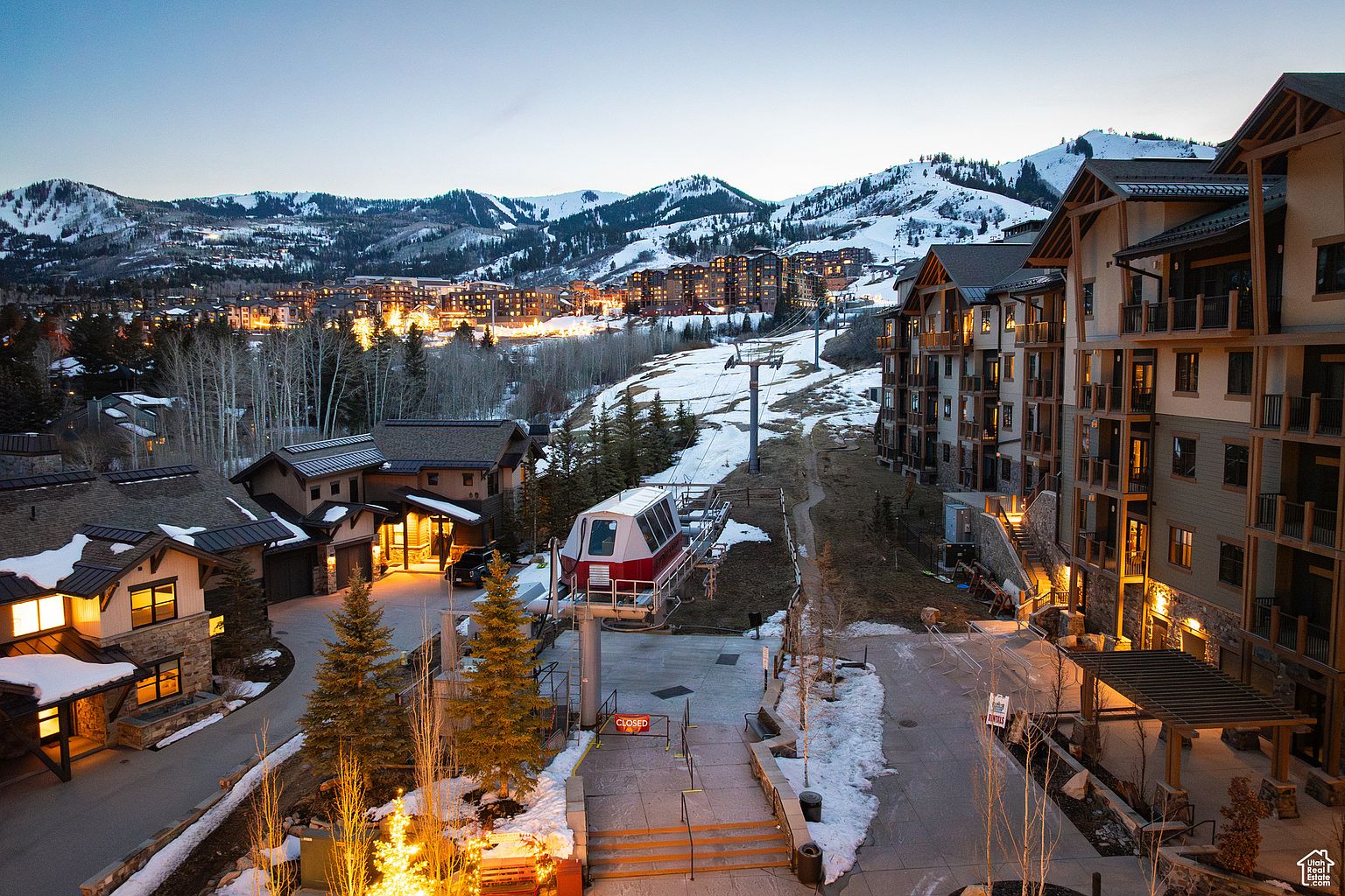 This aerial view showcases a luxurious ski resort at dusk, featuring upscale residences nestled against a backdrop of snow-capped mountains. A ski lift stands prominently in the foreground, connecting the buildings to the slopes. The warm lighting emanating from the buildings creates an inviting atmosphere, highlighting the resort's prime location and amenities.