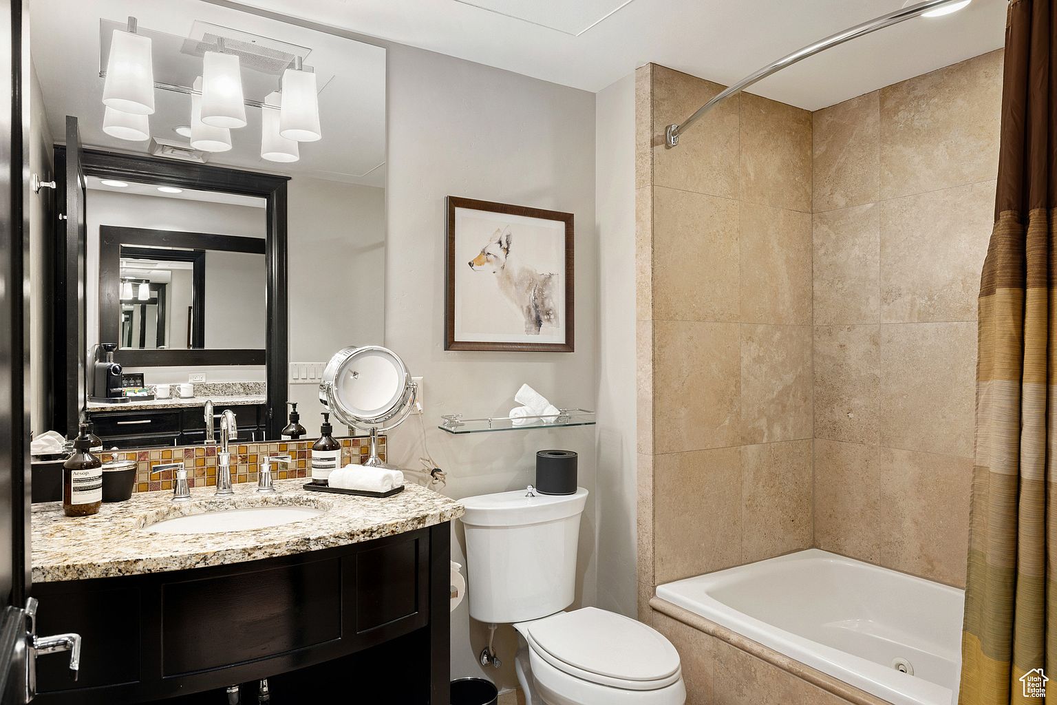 This is a well-appointed primary bathroom featuring a dark wood vanity with a granite countertop and a mosaic tile backsplash. A large mirror reflects the vanity area, and a framed artwork hangs above the toilet. The bathtub is surrounded by beige tiles, and a curved shower rod with a decorative curtain adds a touch of elegance.