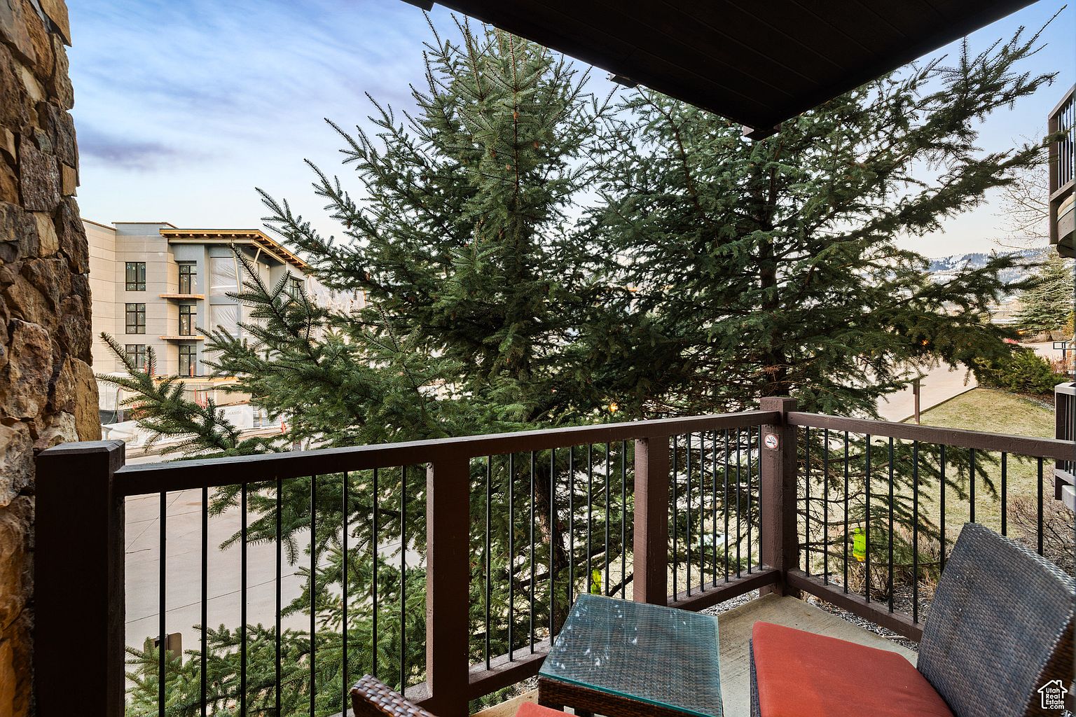 This image showcases a cozy balcony setting with a dark brown railing and a view of lush green trees. A small wicker table and chairs with red cushions suggest a comfortable outdoor seating area. The balcony offers a private and serene space, perfect for relaxation and enjoying the natural surroundings.