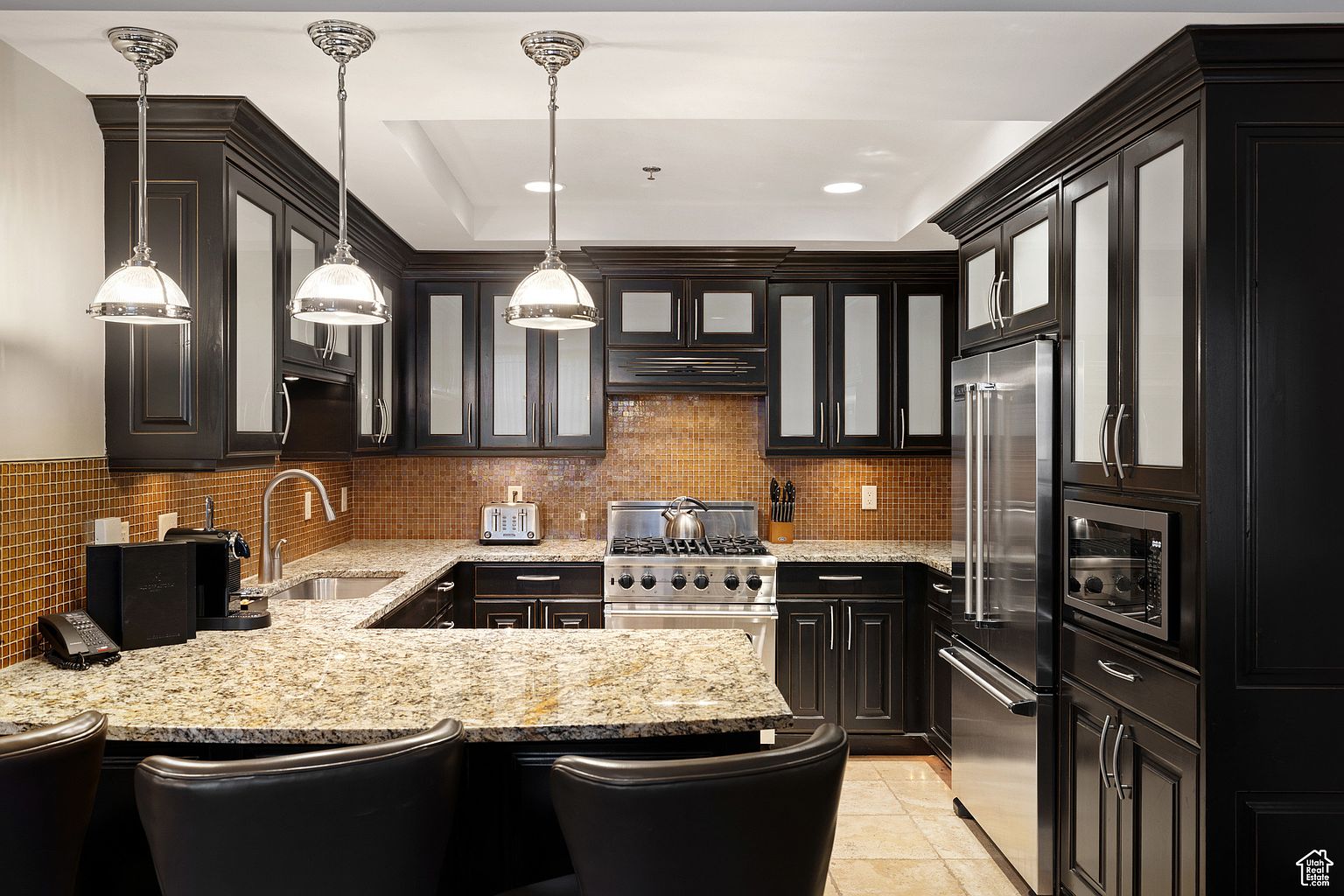 This is a well-appointed kitchen featuring dark wood cabinetry with glass inserts, stainless steel appliances, and granite countertops. The backsplash is a mosaic tile, and pendant lighting hangs over the island. The kitchen has a luxurious and modern feel, perfect for cooking and entertaining.