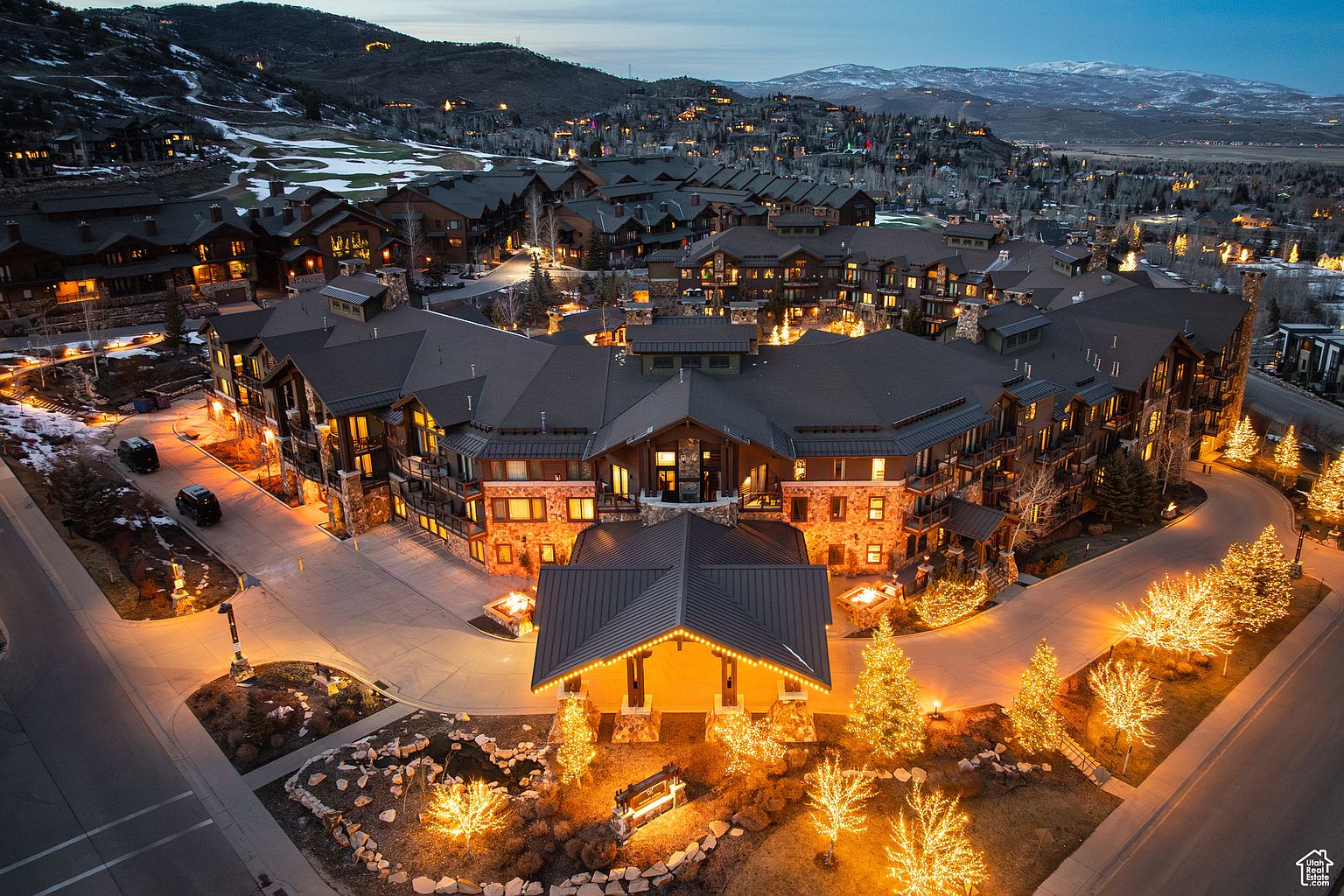 This aerial view showcases a luxurious resort or residential complex at dusk, nestled in a mountainous landscape. The architecture features a blend of stone and wood, with warm lighting emanating from the windows and illuminating the surrounding pathways and landscaping. The scene evokes a sense of upscale living and serene ambiance, highlighting the property's prime location and sophisticated design.