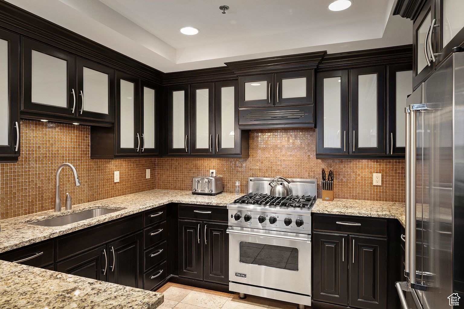 This is a well-lit kitchen featuring dark wood cabinetry with frosted glass panels and stainless steel appliances. The countertops are granite with a tan and brown mosaic tile backsplash. A stainless steel Viking range is the focal point, complemented by a matching refrigerator.