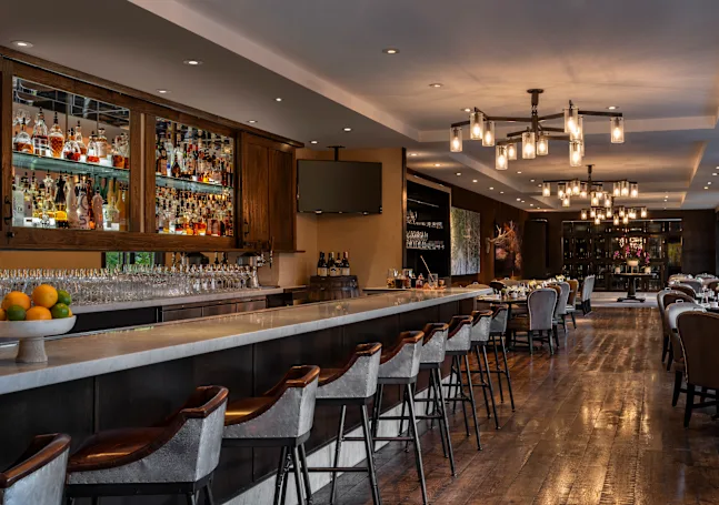 The image showcases a beautifully styled restaurant or dining area with a well-stocked bar featuring a wide selection of liquor. Elegant bar stools line the bar, while tables with upholstered chairs are arranged in the dining area. The lighting consists of modern chandeliers, creating a warm and inviting ambiance.