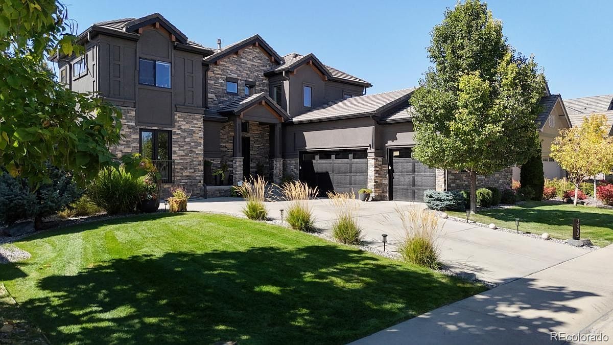 This impressive two-story suburban home features a sophisticated blend of dark stucco and natural stone siding, creating a modern yet warm aesthetic. The property includes a spacious three-car garage, a well-manicured front lawn with ornamental grasses, and a paved driveway leading to a welcoming covered entryway. The bright, clear sky highlights the architectural depth of the rooflines and the inviting curb appeal of this upscale residence.