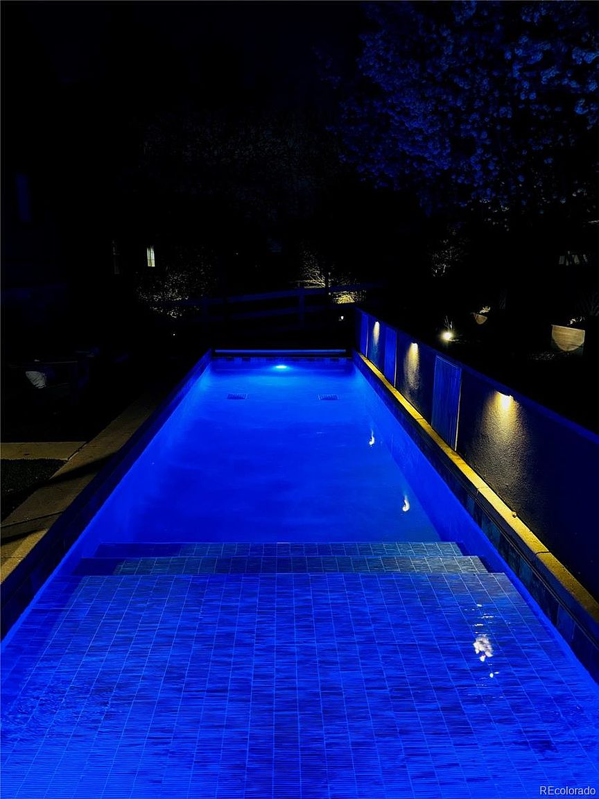This striking night-time shot captures a sleek, rectangular lap pool illuminated by vibrant blue underwater lighting. The pool features a set of wide, tiled steps leading into the water, while warm accent lights mounted on the adjacent wall create a dramatic contrast against the dark surroundings. The perspective is from the shallow end, looking down the length of the pool, emphasizing its modern design and inviting atmosphere.