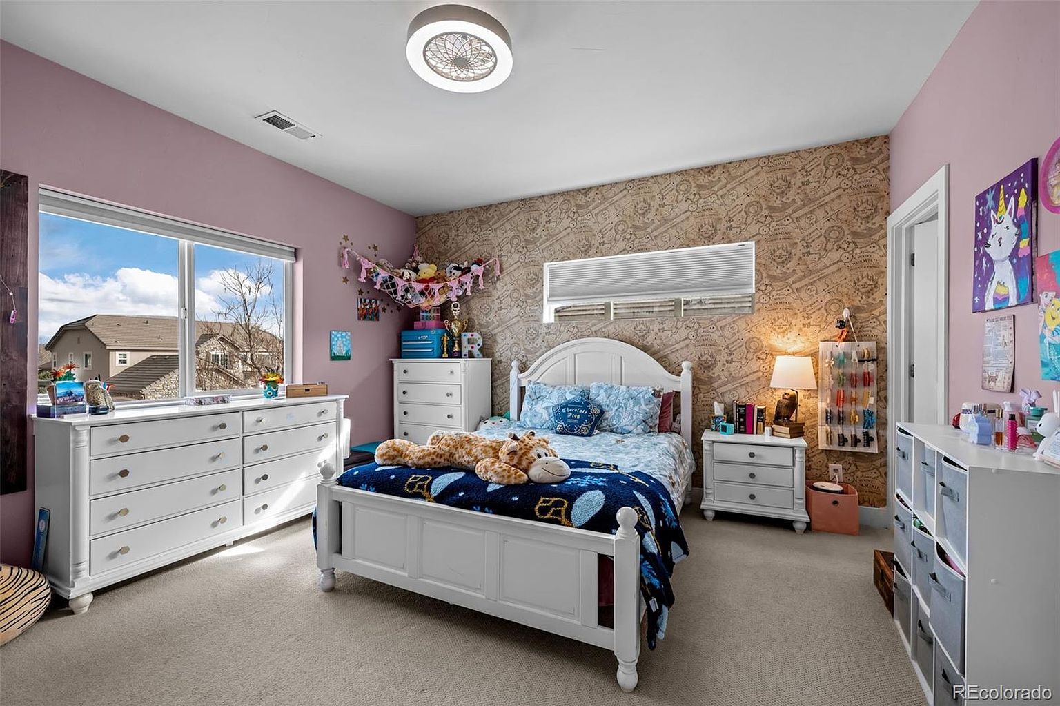 This bright and cheerful bedroom features a white bed frame with a patterned blue quilt and a large plush giraffe toy, set against a unique wallpapered accent wall. The room includes a white dresser, a matching nightstand with a lamp, and a storage unit with bins, all illuminated by a modern ceiling light fixture. Large windows provide ample natural light, creating a welcoming and playful atmosphere suitable for a child or guest.