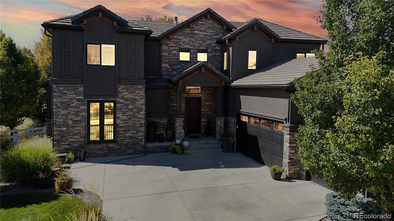 This striking two-story luxury home features a sophisticated blend of dark charcoal stucco and natural stone veneer, creating a modern yet timeless aesthetic. The front facade is highlighted by a prominent entryway, a spacious multi-car garage, and large, well-placed windows that suggest a bright and open interior. The scene is captured at dusk, with the warm glow of interior lighting contrasting beautifully against the dramatic, colorful sunset sky.