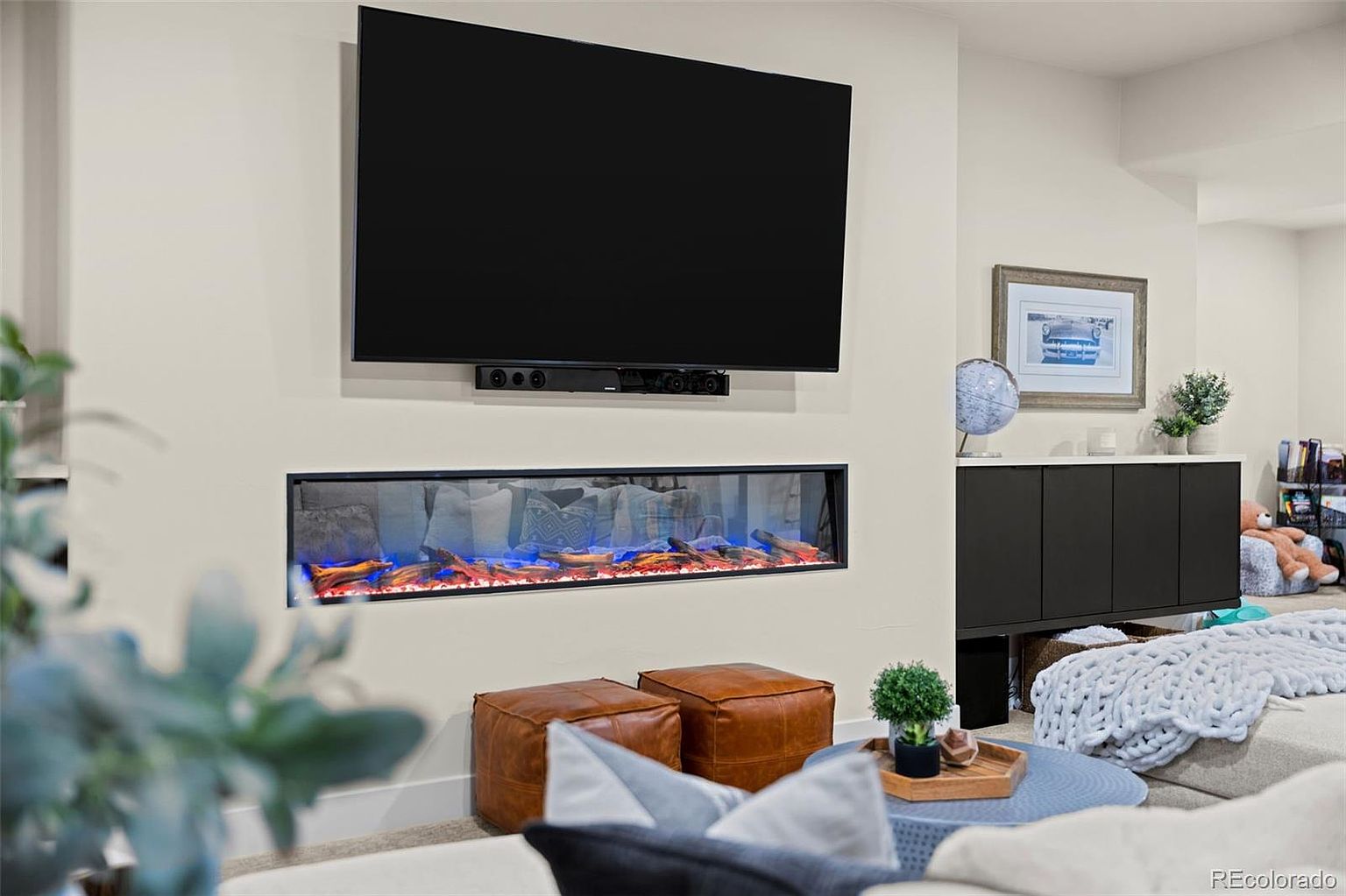 This modern living room features a sleek, linear electric fireplace with glowing embers mounted beneath a large wall-mounted television. The space is accented by two brown leather ottomans, a dark sideboard cabinet, and a cozy, textured throw blanket draped over furniture in the foreground. The neutral color palette and clean lines create a warm, inviting atmosphere perfect for family relaxation.